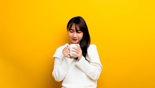 Image of young woman having fun enjoying coffee cup at home smiling beautiful girl drinking hot tea in winter and smiling happily on yellow background.