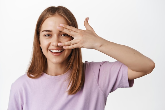 Image of young woman showing her clean face, facial skin without make up, natural feminine look, open fingers near eye and smiling happy, standing against white background