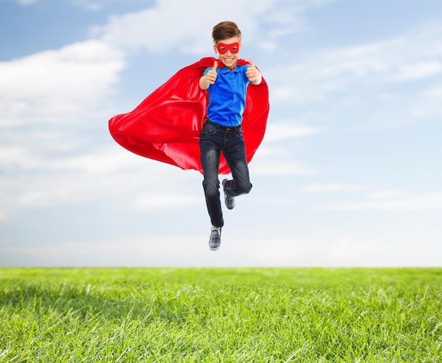 imagination, gesture, childhood, movement and people concept - boy in red super hero cape and mask flying in air and showing thumbs up over blue sky and grass background