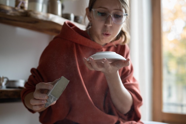 Immersive in process girl blowing on porcelain plate to remove excess dust sitting in craft workshop