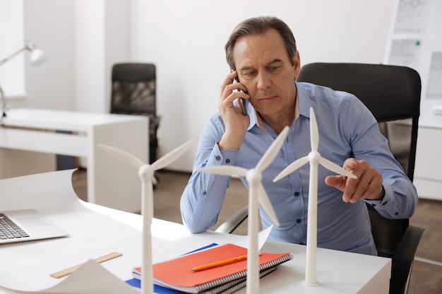 Important call. Serious professional engineer sitting at the table and talking on smart phone while working on constructing eco turbines