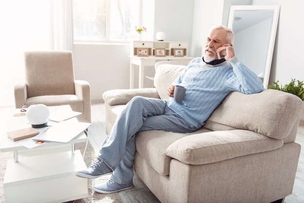 Important discussion. Pleasant senior man sitting on the couch in the living room and having a serious conversation on the phone while drinking coffee