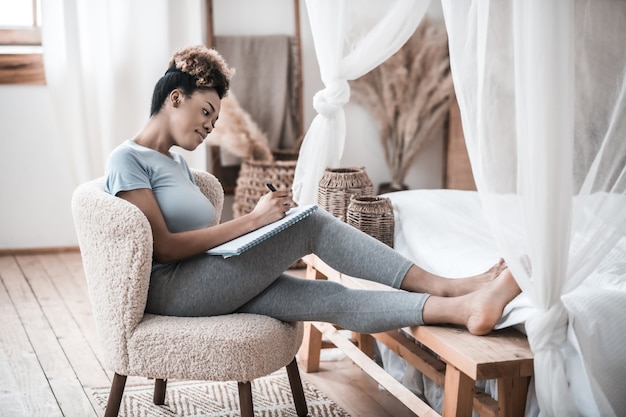 Important occupation. Young adult dark skinned woman sitting in chair with her feet on bed writing notes in notebook