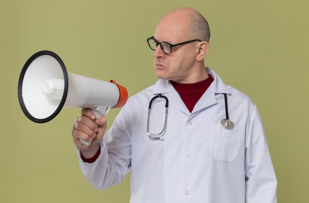 Impressed adult slavic man with optical glasses in doctor uniform with stethoscope holding and looking at loud speaker 