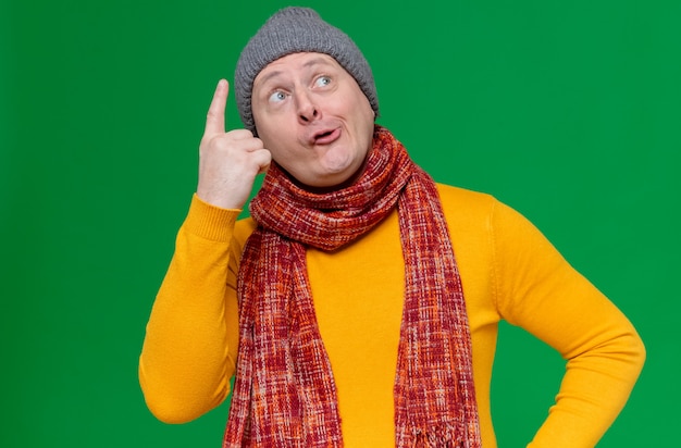 Impressed adult slavic man with winter hat and scarf around his neck pointing up 