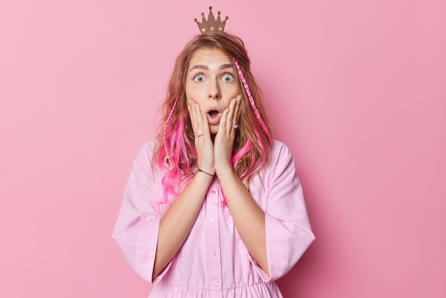 Impressed blue eyed European woman princess keeps hands on cheeks mouth opened stares with excitement and shock wears small crown on head and shirt isolated over pink background. Omg concept