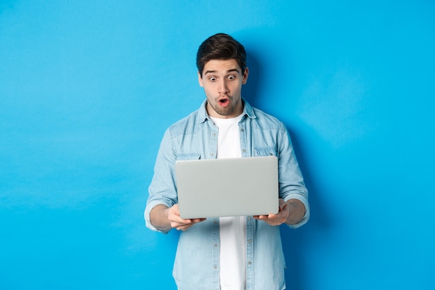 Impressed caucasian guy looking at laptop screen with amazement, checking out promo in internet, standing against blue background