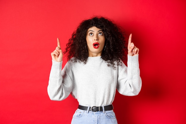 Impressed european female model in casual clothes, checking out promo with dropped jaw, looking and pointing fingers up, showing logo, standing on red background.