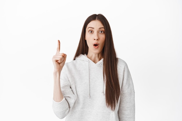 Impressed girl telling about promotional offer pointing finger up gasping and looking shocked at camera say wow recommending follow website click on banner white background