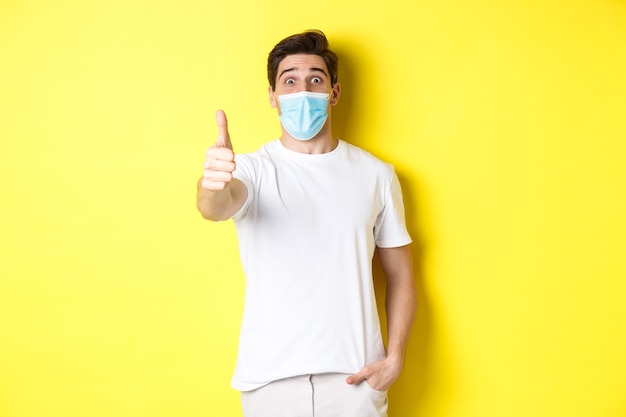 Impressed guy in medical mask showing thumb up in approval, like something awesome, yellow wall