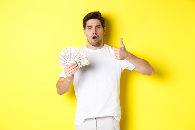 Impressed man showing thumb up, holding money credit, standing over yellow wall