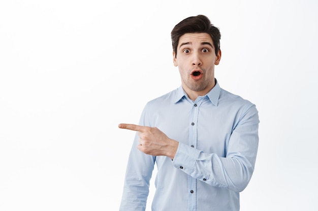 Impressed office worker, manager in suit, gasping amazed, pointing finger left at promo offer, showing advertisement with excited face, standing over white wall