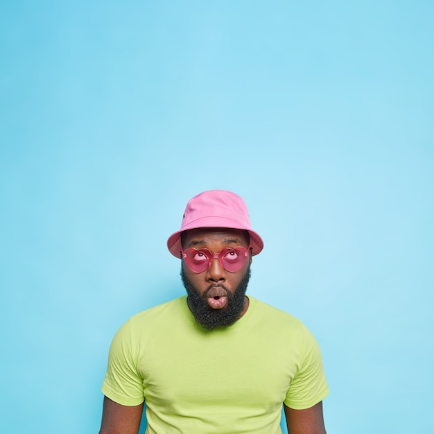 Impressed surprised guy with thick beard focused above keeps mouth opened wears panama casaul green t shirt heart shaped sunglasses isolated over blue wall