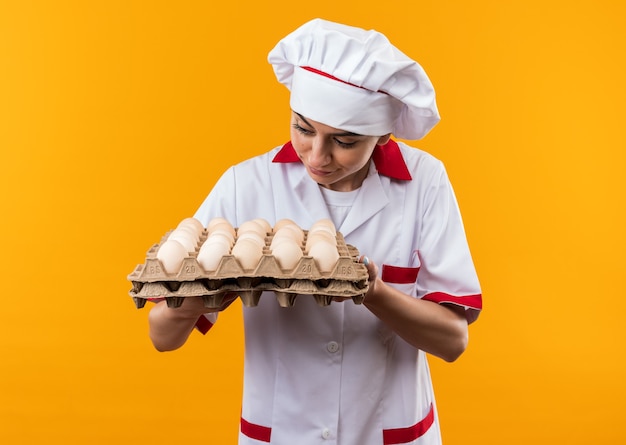 Impressed young beautiful girl in chef uniform holding and looking at batch of eggs 