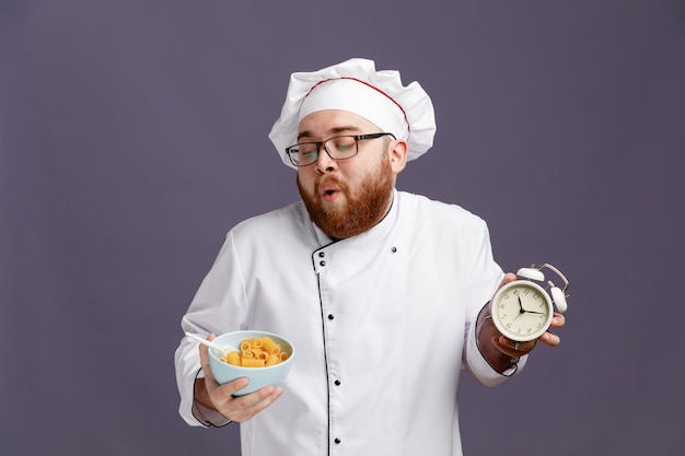 Impressed young chef wearing glasses uniform and cap holding alarm clock and bowl of macaroni pasta with spoon in it looking at bowl isolated on purple background