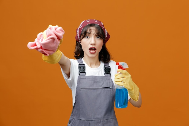 Impressed young female cleaner wearing uniform rubber gloves and bandana holding cleanser stretching cloth duster out towards camera looking at camera isolated on orange background