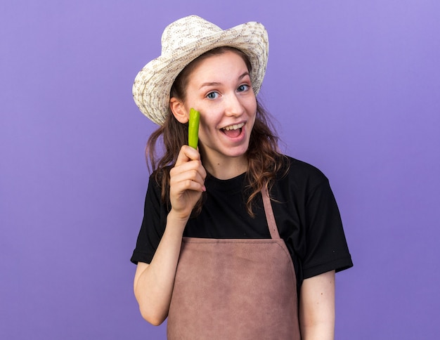 Impressed young female gardener wearing gardening hat holding pepper 