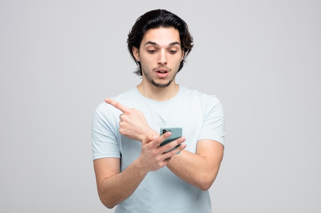 impressed young handsome man holding and looking at mobile phone pointing to side isolated on white background