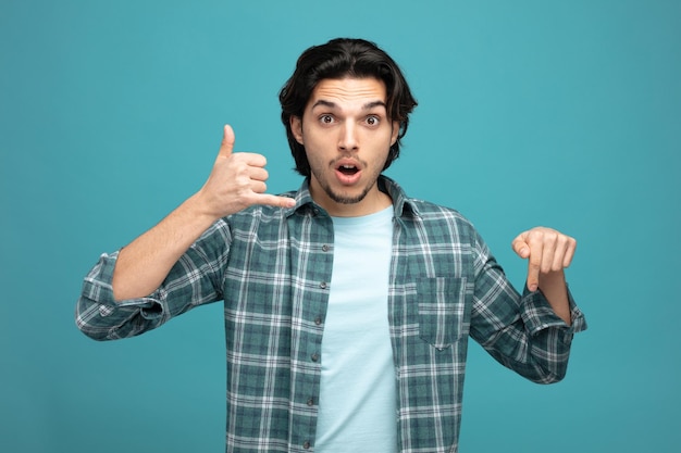 impressed young handsome man looking at camera showing call gesture pointing down isolated on blue background