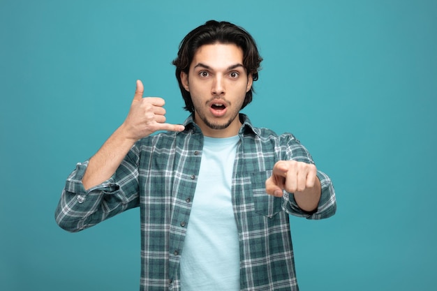 impressed young handsome man looking and pointing at camera showing call gesture isolated on blue background