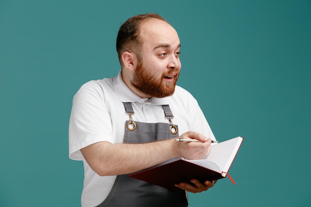 Impressed young male barber wearing white shirt and barber apron holding pen and note pad looking at side isolated on blue background