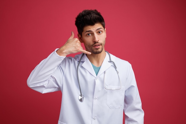 Impressed young male doctor wearing medical uniform and stethoscope around his neck looking at camera showing call me gesture isolated on red background