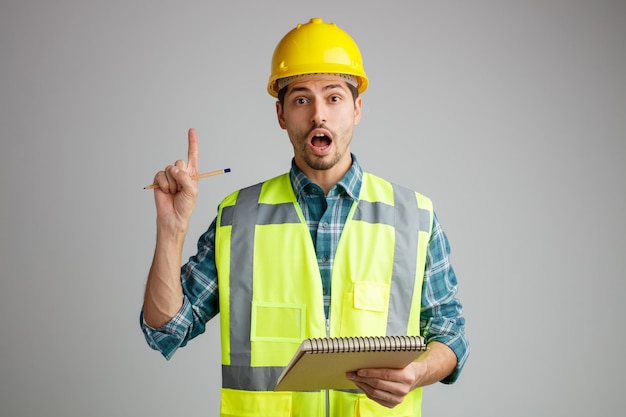 Impressed young male engineer wearing safety helmet and uniform holding note pad and pencil looking at camera pointing up isolated on white background