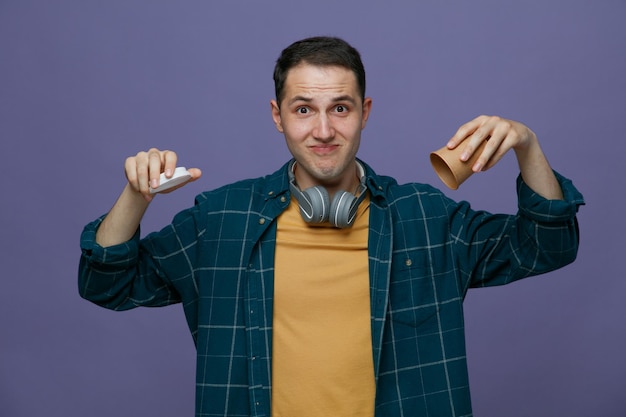 Impressed young male student wearing headphones around neck looking at camera holding paper coffee cup upside down and its cap isolated on purple background