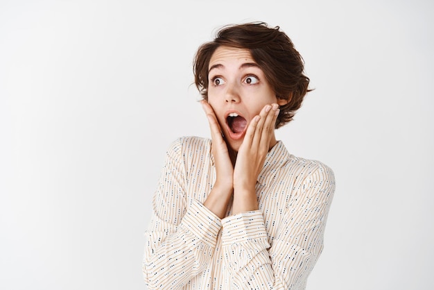 Impressed young woman in blouse drop jaw gasping and look fascinated at empty space staring left and touching cheeks from amazement standing on white background