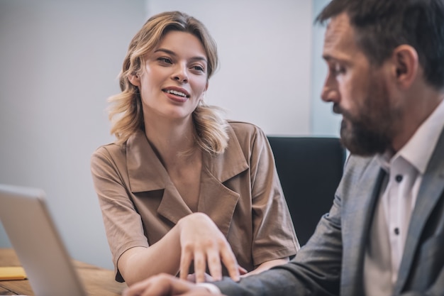 Increased interest. Cheerful young blonde beautiful woman touching hands of serious male colleague in business suit sitting in office