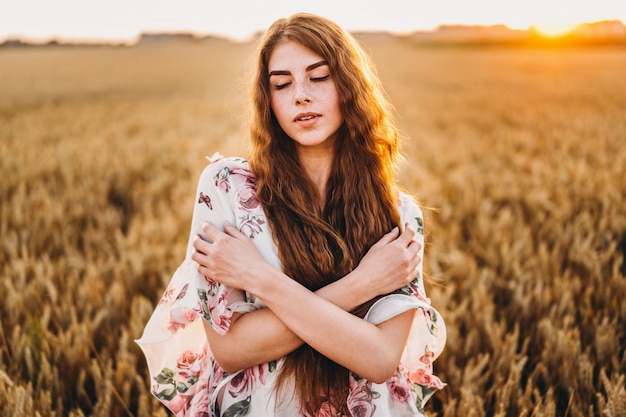 Incredible young woman with long curly hair and freckles face. Woman in dress posing in wheat field at sunset. Close up portrait