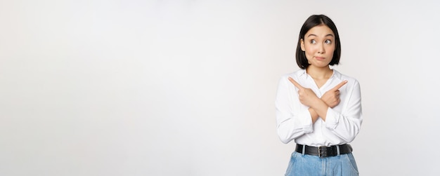 Indecisive asian woman pointing fingers sideways pointing fingers and looking clueless confused with choices standing over white background