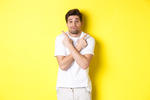 Indecisive man pointing fingers sideways, struggling to make decision, asking advice, standing over yellow wall