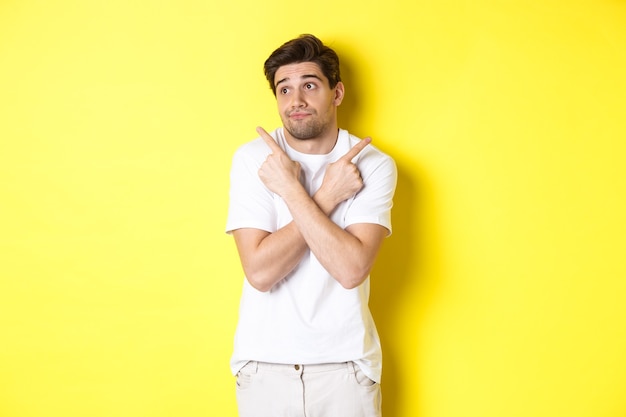 Indecisive man shrugging, pointing fingers sideways, troubled to choose, standing over yellow wall