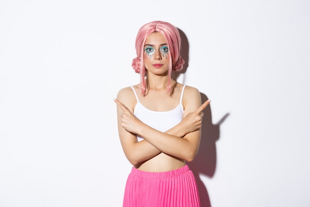 Indecisive young woman in pink wig and halloween costume, pointing sideways at two choices, showing variants, standing over white background