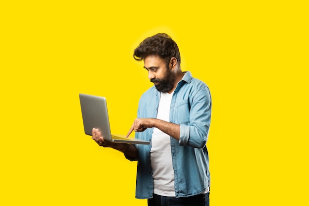 Indian asian bearded young man in casual cloths using laptop while standing on yellow