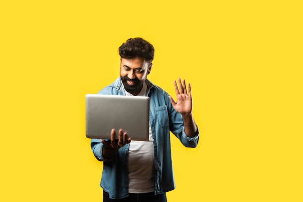 Indian asian bearded young man in casual cloths using laptop while standing on yellow