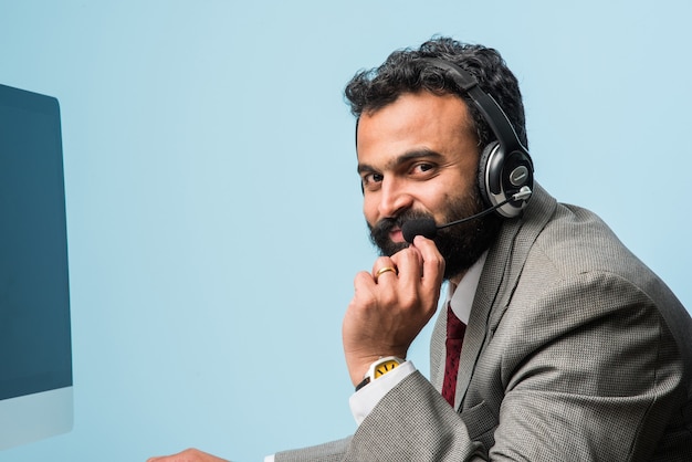 Photo indian asian bearded young man in suit in call centre, indoor, listening to headphones, browsing  computer or having a voice call