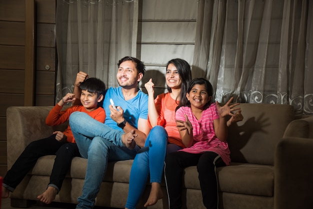 Indian asian family of four watching television or TV while sitting on sofa or couch