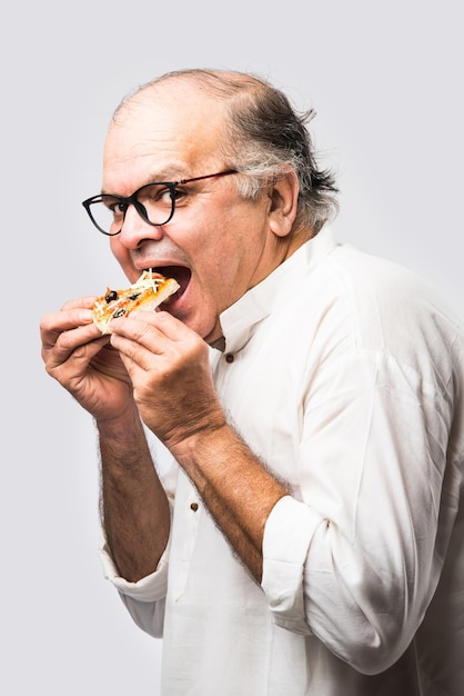 Indian asian senior or old man eating pizza while standing isolated against white wall