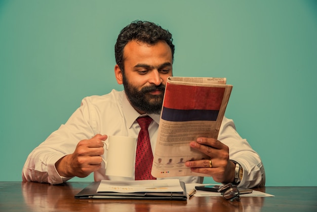 Indian asian young businessman reading newspaper while having coffee at office desk