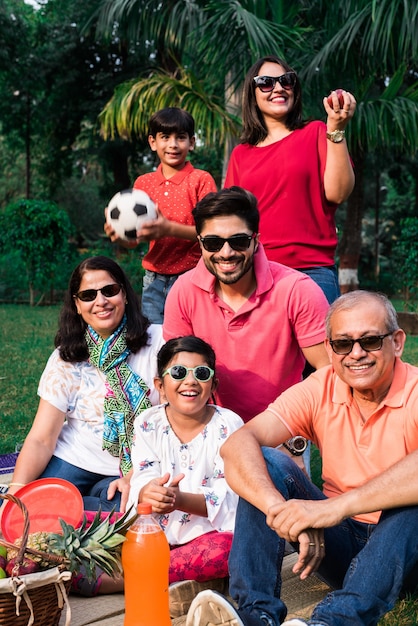 Indian Family enjoying Picnic - Multi generation of asian family sitting over lawn or green grass in park with fruit basket, mat and drinks. selective focus