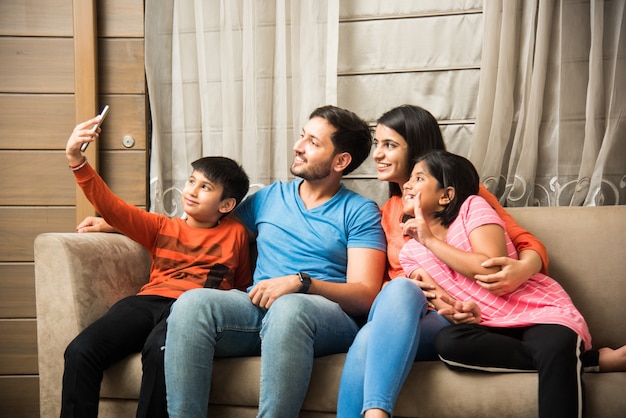 Indian family sitting on sofa and using smartphone laptop or tablet watching movie or surfing internet