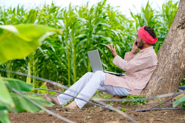 Indian farmer using laptop and talking on smartphone at agriculture field.