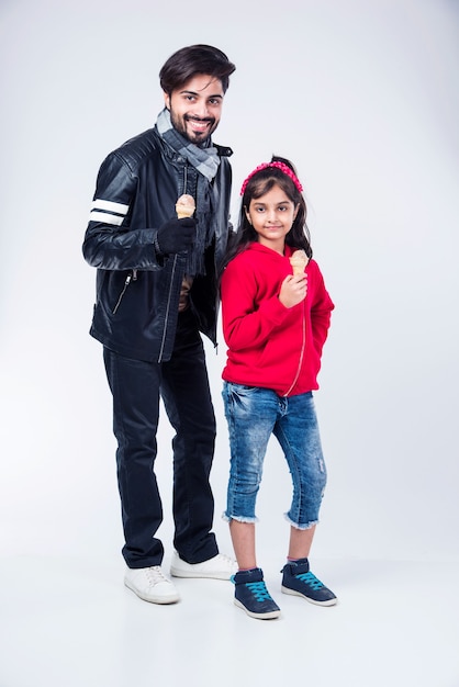 Indian father and daughter eating ice cream in cone while wearing warm clothes on white background