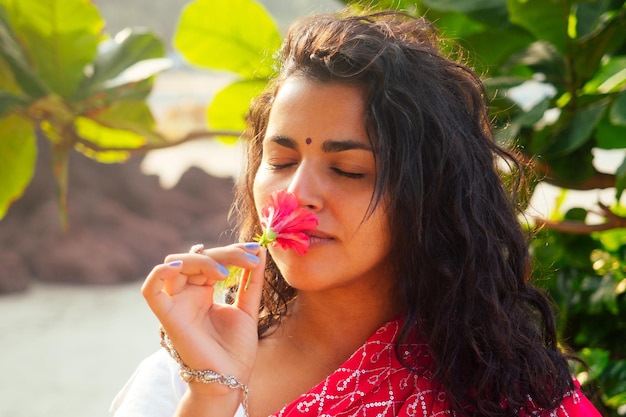 Indian female fashion model white-toothed smile with flower in her curly hair in traditional india outfit red wedding sari posing near tropical trees on a paradise beach sea ocean.wellness spa resort