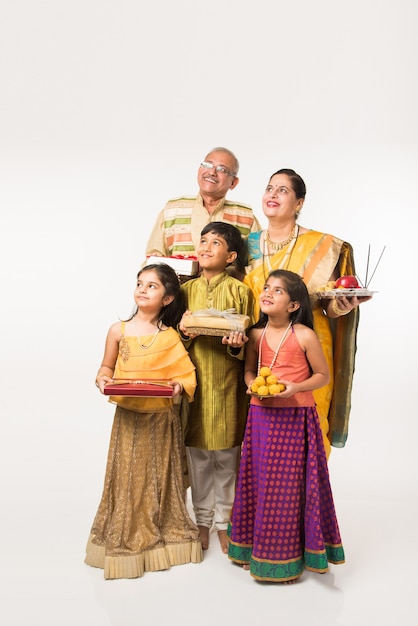 Indian kids with grandparents in traditional wear holding gifts, sweets and puja or pooja thali or taking selfie, isolated over white background