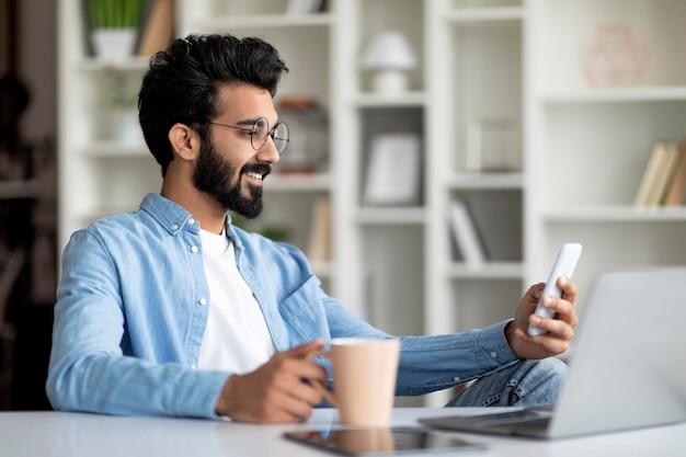 Indian male freelancer using smartphone and drinking coffee at home office