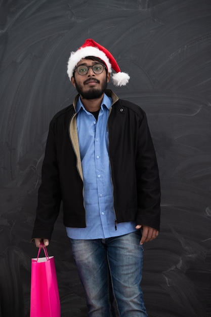 Photo indian  man wearing party clothes in santa hat with shopping bag on dark  background studio dark skinned middle eastern santa claus merry christmas