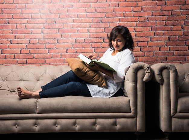 Indian mid age lady or woman reading book while sitting over couch or sofa, having coffee